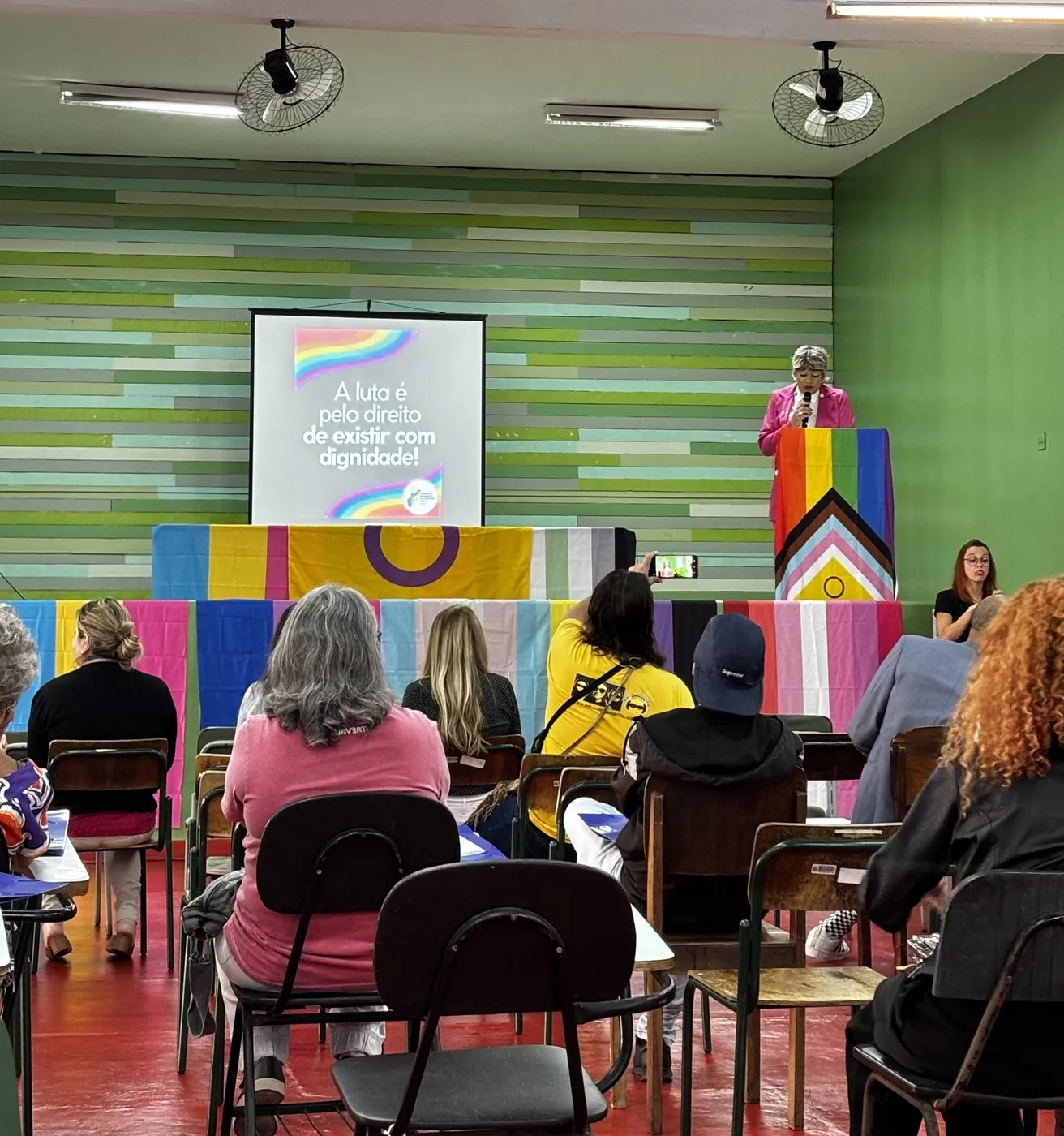 Auditório da 1ª Conferência LGBTQIA+ de Marília, com bandeiras e o telão: A luta é pelo direito de existir com dignidade