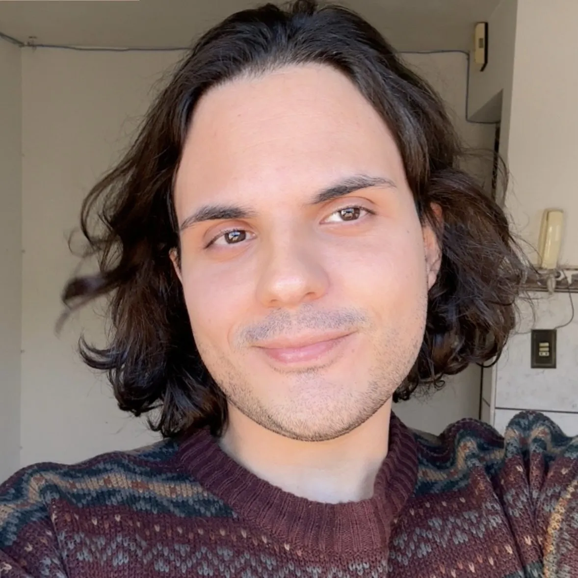 Gabriel Meyer, psicólogo, sorrindo, de cabelo comprido e suéter vinho